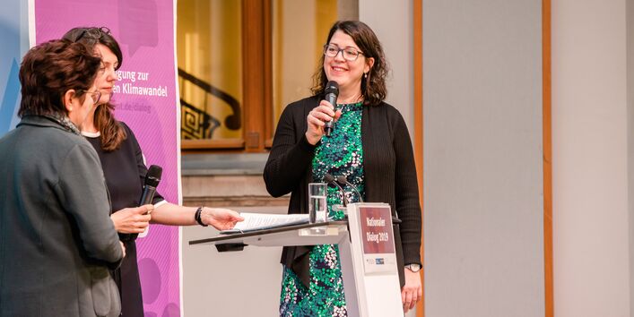 Drei Vortragende stehen am Stehpult und halten Mikrofone in den Händen. Sie lachen sich gegenseitig an.Im Hintergrund steht das lilafarbene Veranstaltungs-Rollup