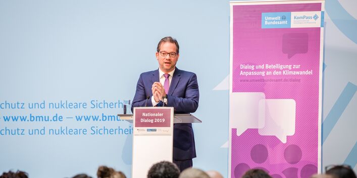 Redner steht am Stehpult und trägt vor. Aufsteller im Hintergrund sowie vorne auf dem Stehpult tragen vorwiegend in lila gehalten den Veranstaltungstitel