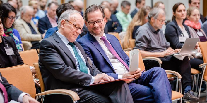 Teilnehmende sitzen auf Stühlen in Reihen und beschäftigen sich mit Schriftgut in ihren Händen.