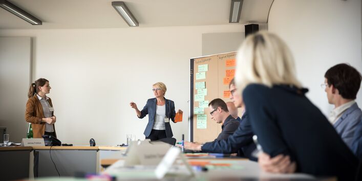 Vortragende steht an mit Moderationskarten bestückte Moderationswand und hält beschriftete orangene Moderationskarte in der Hand und spricht in den Raum, wo andere Teilnehmerinnen und Teilnehmer am Tisch in Reihe sitzend zuhören. Im Hintergrund steht eine weitere Teilnehmerin.