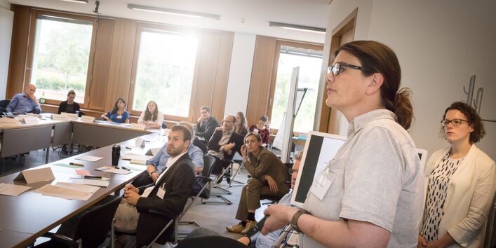 Vortragende hält die Fernbedienung zur Präsentation in der Hand und steht seitlich zur Präsentationswand zusammen mit einer anderen Teilnehmerin und schauen wie die anderen in Richtung Präsentationswand. Die anderen Teilnehmerinnen und Teilnehmer sitzen sich in Reihe an Tischen gegenüber oder einzeln auf Stühlen.