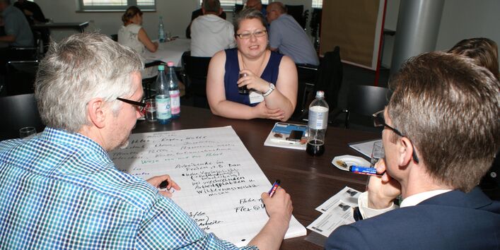 Teilnehmer sitzen im Arbeitsgruppenraum am Arbeitsgruppentisch und diskutieren miteinander.