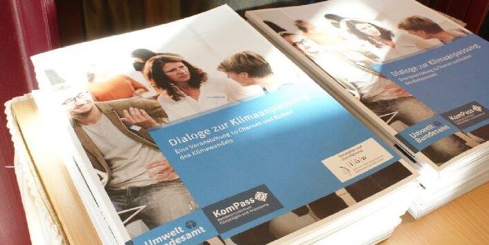 Das Bild zeigt zwei Stapel Broschüren mit dem Titel Dialoge zur Klimaanpassung. Eine Veranstaltung zur Chancen und Risiken des Klimawandels. Der Buchdeckel zeigt Menschen im Dialog. Eine Frau gibt dem Gesagten Ausdruck mit den Händen. Ein Mann nickt zustimmend im Hintergrund. Eine andere Frau hört zu.