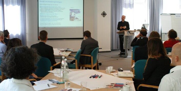 Vortragender steht am Rednerpult vor den Teilnehmern, die an den Arbeitsgruppentischen zuhörend sitzen. Auf den Tischen liegen Moderationsmaterialien wie Stifte, Moderationskarten.