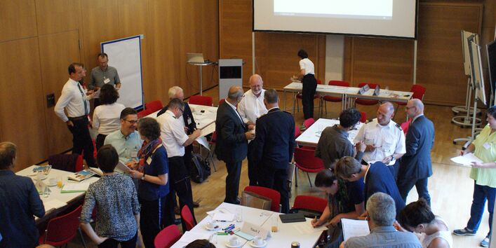 Teilnehmende stehen miteinander unterhaltend in lockeren Gesprächsgruppen beieinander in dem holzvertäfelten Veranstaltungsraum. Im Hintergrund stehen Rednerpult und die große Leinwand. Auf den Tischen befinden sich Arbeitsmaterialien und Getränke.