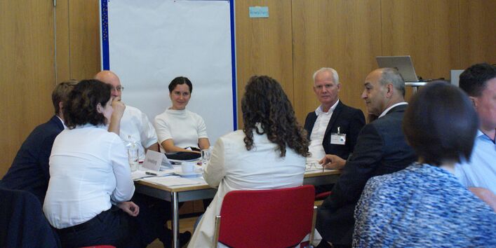 Teilnehmende sitzen an einem Gruppenarbeitstisch und hören einer Teilnehmerin zu. Im Hintergrund steht eine Pinwand mit einem großen leeren weißen Blatt bespannt. 