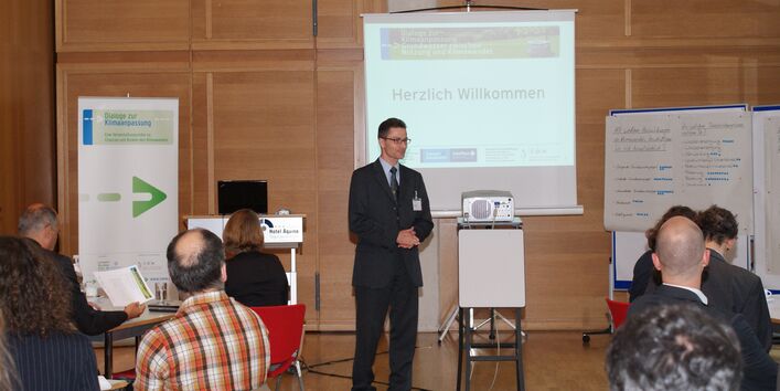 Vortragender begrüßt die Teilnehmer. Seine Hände sind gefalten. Im Hintergrund stehen Moderationswände, der Veranstaltungsaufsteller Dialoge zur Klimaanpassung und eine Leinwand mit dem Schriftzug Herzlich Willkommen.