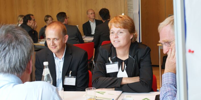 Zwei Teilnehmende sitzen nebeneinander am Gruppenarbeitstisch und hören anderen Teilnehmern zu. Im Hintergrund sitzen andere Teilnehmer am anderen Gruppenarbeitisch. Neben diesem Gruppenarbeitstisch steht eine Moderationswand.