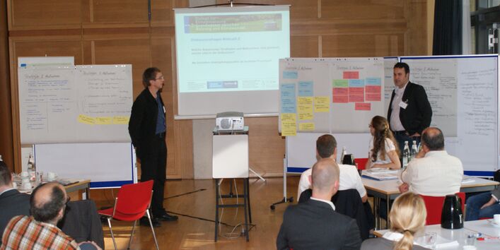 Zwei Teilnehmer präsentieren Ergebnisse an Moderationswänden. An den Moderationswänden hängen farbige Moderationskarten. Im Hintergrund sieht man noch eine Präsenation auf der Leinwand. Im Vordergrund sitzen Zuhörer an den Gruppenarbeitstischen.