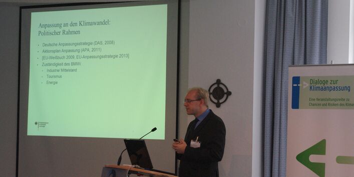 Vortragender steht am Rednerpult. Im Hintergrund ist die Präsentation auf Projektionswand zu sehen und der Veranstaltungsaufsteller.