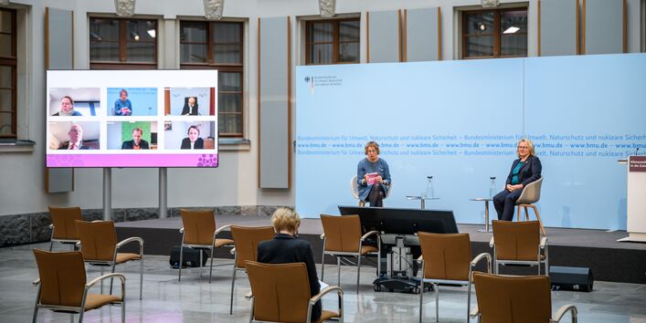 In dem hellen historischen Innenhof sitzen 2 Frauen auf Schalenstühlen auf dem Podium vor einem blauem Hintergrund. Links davon steht ein großer flacher Bildschirm, der die zugeschalteten Konferenzteilnehmenden zeigt. Vor dem Podium stehen leere Stühle. Auf einem Stuhl beugt sich eine Teilnehmerin über Ihre Unterlagen.