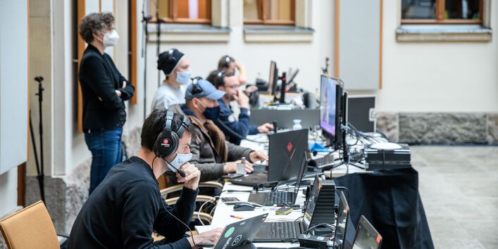 Die Technikerinnen und Techniker sitzen in einer langen Reihe und schauen konzentriert in die Laptops. Sie tragen Kopfhörer und Mund-/Nasenbedeckungen.