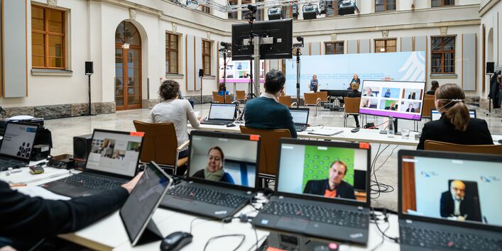In dem hellen historischen Innenhof geht der Blick über den langen Tisch mit dem Laptops hinüber über sitzendes Publikum zum Podium. Auf dem Podium sitzen entfernt zwei Menschen vor blauem Hintergrund. Im Hof stehen verteilt auf Ständern die flachen Bildschirme, die die dazugeschalteten Teilnehmenden zeigen. Kamera- und Lichttechnik hängt  in der Höhe seitlich entlang der Fassade an Metallstreben.