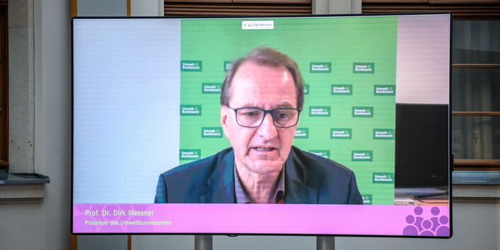 Auf einem flachen Bildschirm im hellen historischen Innenhof ist im Großformat zu sehen wie der Präsident des Umweltbundesamts einen Beitrag spricht.