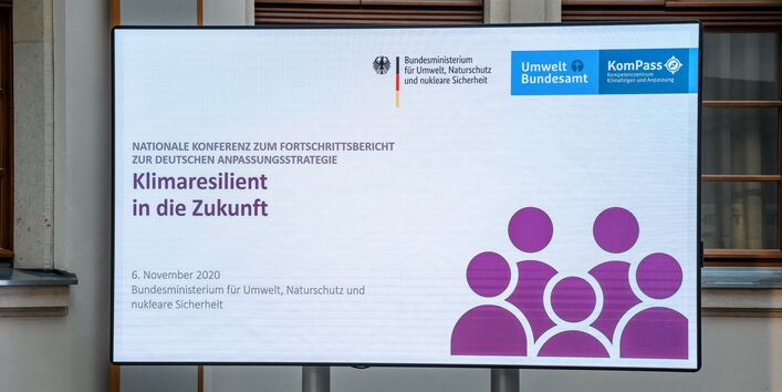 Der Blick geht auf das Konferenzlogo, welches sich im hellen historischen Innenhof auf dem großen flachen Bildschirm nahe zeigt