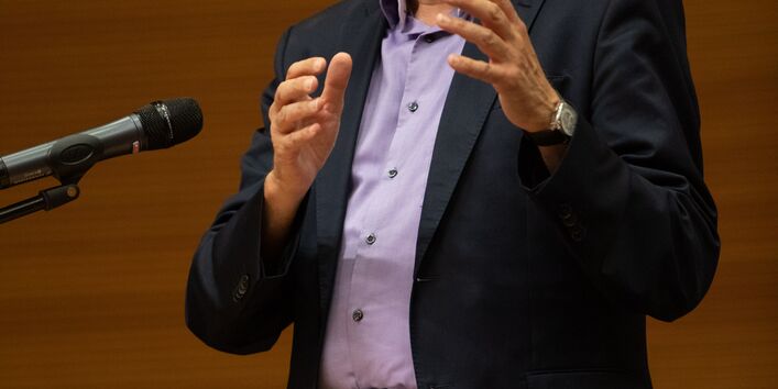 Ein Vortragender spricht gestikulierend ins Mikrofon. Er steht an einem Rednerpult und trägt einen dunklen Anzug mit einem zartlila Hemd unter dem Jacket.