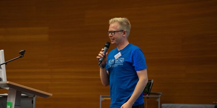 Ein Vortragender spricht in ein Mikrofon, welches er in der Hand hält. Der Vortragende trägt ein blaufarbenes T-Shirt und eine Brille.