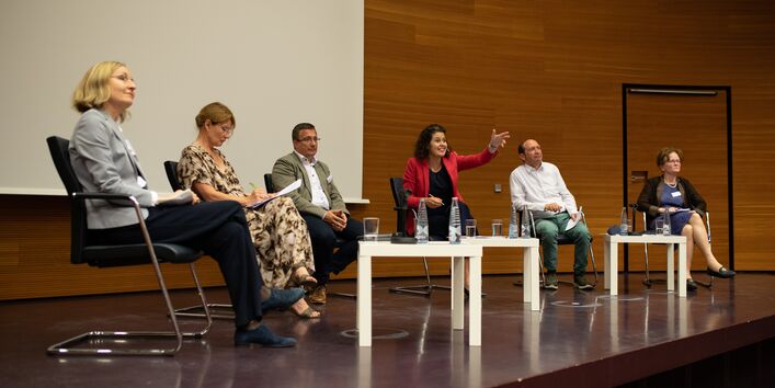 6 Podiumsteilnehmende sitzen auf der Bühne im Halbkreis vor hellen niedrigen Tischen und tauschen sich gestikulierend mit dem Publikum aus. Eine Frau im roten Sakko zeigt mit gestrecktem Arm in Richtung Publikum.