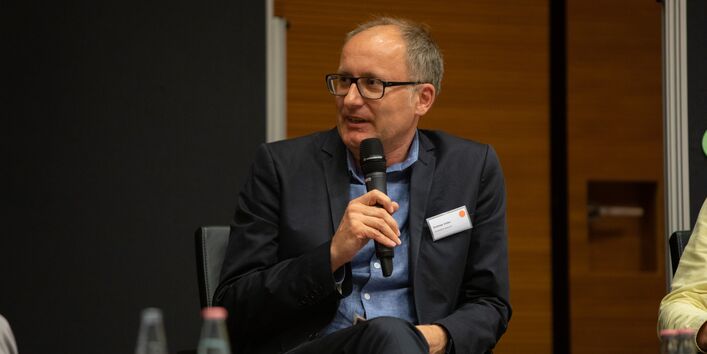Ein Teilnehmender sitzt auf dem Podium und spricht in das Mikrofon in Richtung der anderen Podiumsteilnehmenden. Er hält das Mikrofon in der Hand. Er trägt ein Namensschild am dunklen Revers.