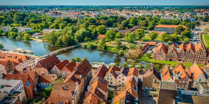 Der Blick geht von oben in die Ferne über eine Stadt mit roten Ziegelsteindächern. Die Stadt ist umgeben von Grün und liegt an einem Fluss. Der Himmel ist blau.