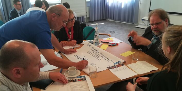 Teilnehmender beugt sich über den Gruppenarbeitstisch und schreibt auf Papier mit was ein Mitteilnehmender spricht.