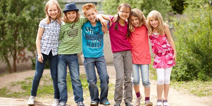 6 Kinder stehen lachend auf einem liegenden Baumstamm für ein Gruppenfoto, die Arme jeweils auf den Schultern des Nachbarn