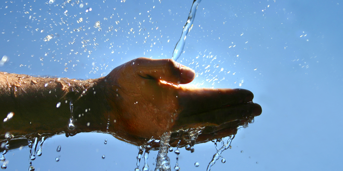 Zwei Hände werden in einen Wasserstrahl gehalten. Im Hintergrund scheint die Sonne