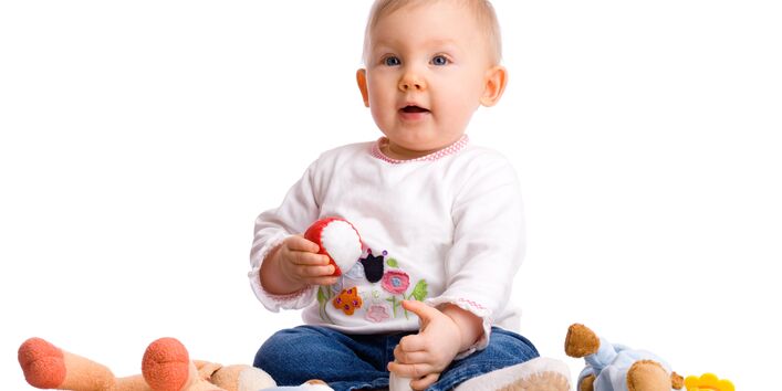 Ein Baby sitzt neben Kuscheltieren, es hält einen Ball in der Hand und guckt erwartungsvoll nach oben.