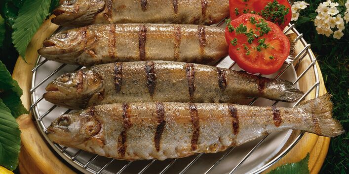 3 gegrillte Fische serviert auf einem Grill umgeben von einigen Blumen und garniert mit 2 halben Tomaten
