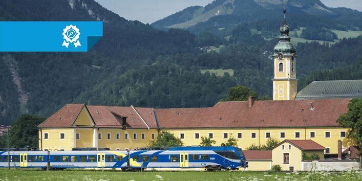 Im Vordergrund fährt ein Personenzug vor einem größen, gelb gestrichenen Gebäude vorbei. Zu dem Gebäude gehört ein Turm, der rechts im Bild zu sehen ist. Im Hintergrund sind Berge erkennbar. Links oben über dem Bild liegt ein Banner des Blauen Kompasses.