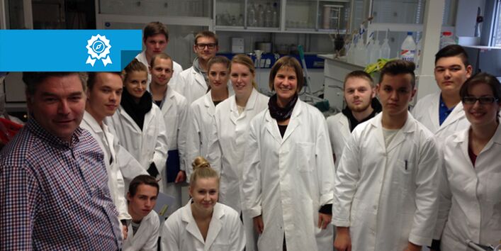 Gruppenbild aus dem Labor. Personen tragen weiße Kittel. Im Hintergrund befinden sich Labormaterilien in und außerhalb von verglasten Laborschränke. Eine Mann im linken Vordergrund trägt Alltagskleidung: Hose und Hemd.