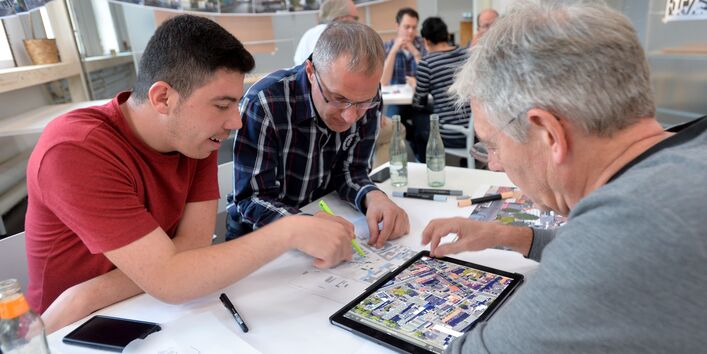 3 Männer sitzen an einem Arbeitsgruppentisch und betrachten Skizzen auf einem Tablet und Papier. Der Mann in einem roten T-Shirt zeigt etwas auf der Papierskizze. Im Hintergrund befindet sich ein weiterer Arbeitsgruppentisch. Bilder sind im Wäscheleinenformat im Hintergrund aufgehängt.
