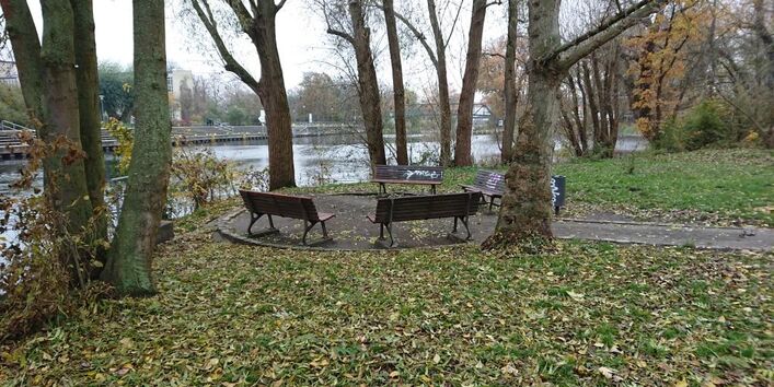 Parkbänke stehen kreisförmig zusammen unter unbelaubten Bäumen am Wasser. Im Hintergrund sind Bäume, Häuser und eine Brücke zu sehen.