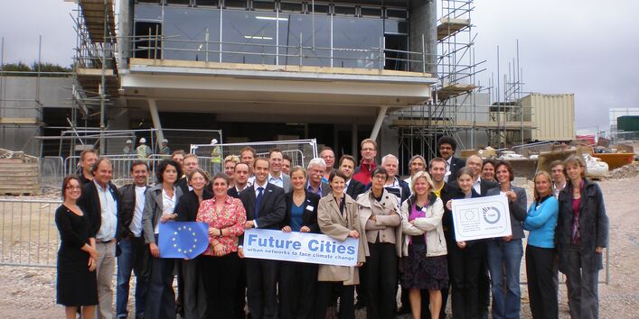 Viele Personen vor einem Haus im Bau, die die EU-Fahne und das Future Cities-Logo hochhalten