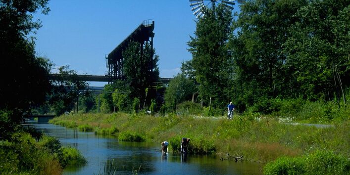 Ein Gewässer in einem Landschaftspark. Im Hintergrund steht ein Windrad. Im Vordergrund spielen Kinder in den Uferbereichen und fangen Fische.