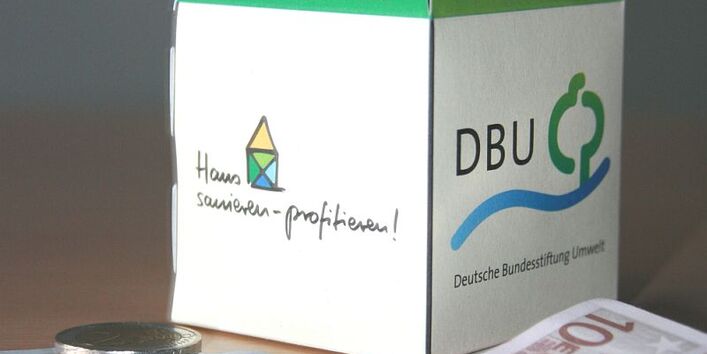 Ein aus Papier gefaltetes Haus mit der Aufschrift DBU steht auf einem Tisch, davor liegt Geld