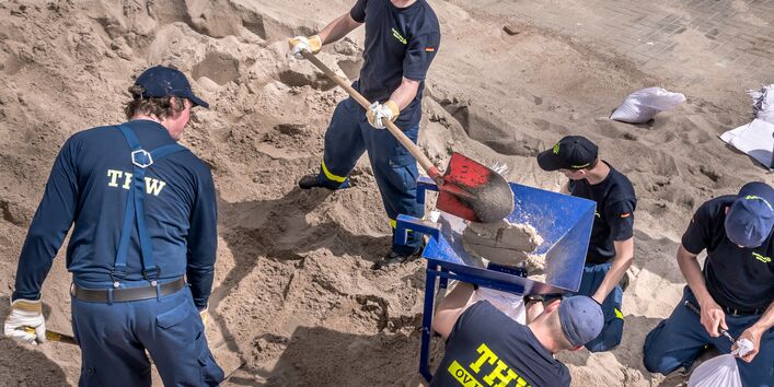 Ausbildungsteilnehmer_innen in Blau gekleidet und mit gelbem Schriftzug THW befüllen Säcke mit Sand mit Hilfe eines roten Spatens und einer blauen Schubkarre.