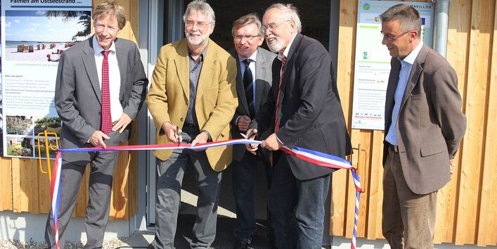 Personen stehen vor einem Strandpavillon, eine Person durchtrennt ein blau-rot-weißes Band mit der Schere