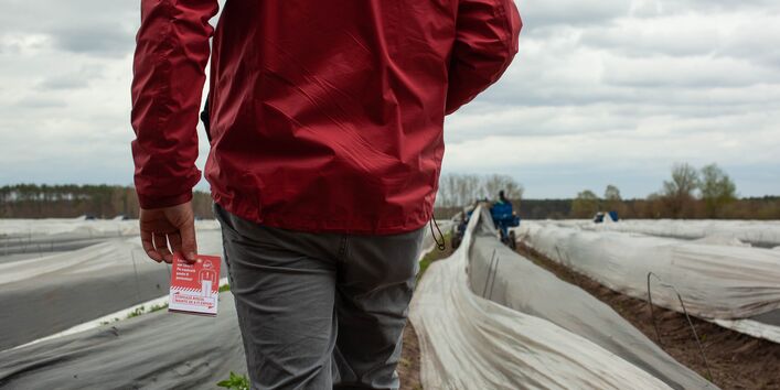 Fotografie einer Person auf einem bewirtschafteten Feld. Nur der Rumpf ist erkennbar, in der Hand hält die Person einen Flyer.