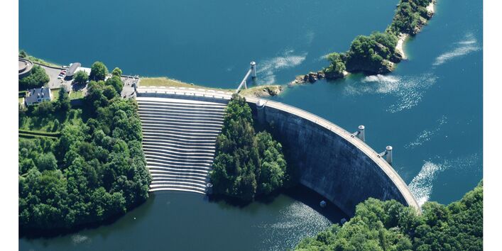 Blick von oben auf eine Talsperre. Treppen führen hinab auf der Seite mit niedrigerer Wasserhöhe. Grün belaubte Bäume auf einem Wall im Wasser und drum herum sowie Gebäudeteile auf der Talsperre runden das Bild ab. Das Wasser erscheint in einem tiefen klaren Blau.
