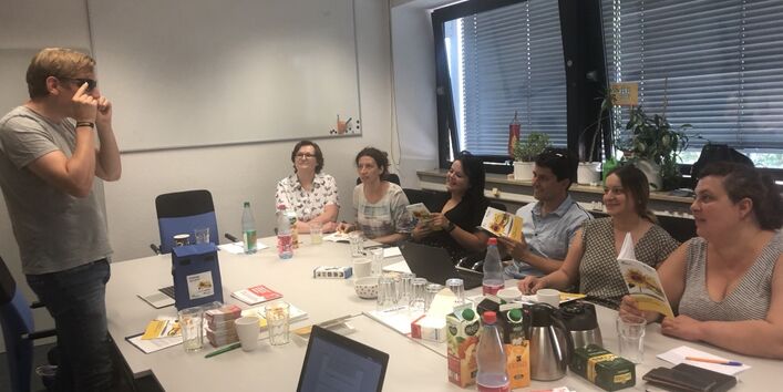 An einem Tisch in einem Schulungsraum sitzen mehrere Personen auf einer Seite, ihnen gegenübersteht eine weitere Person, die etwas erklärt.