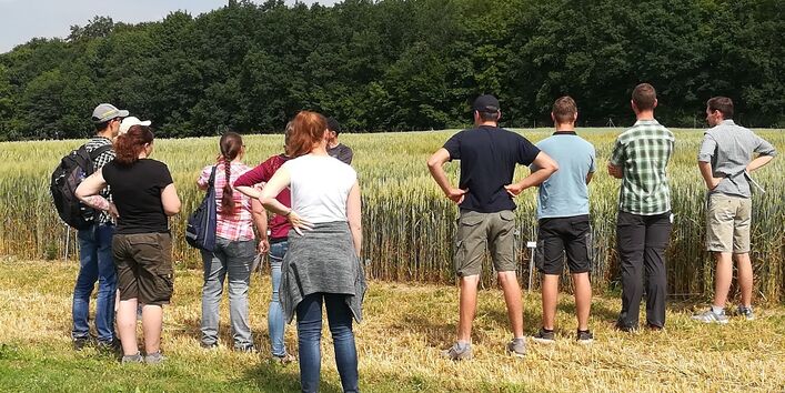Auf einem Acker stehen mehrere jüngere Personen mit dem Rücken zur Kamera. Eine verdeckte Person hält einen Vortrag, dem die anderen zuhören.