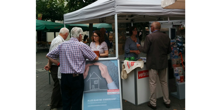 Mehrere Personen stehen um und hinter einem Marktstand der Verbraucherzentrale NRW mit heller Überdachung. Im Vordergrund ein Aufstellplakat, auf dem sich schützend eine Hand über ein mit Kreide gezeichnetes Haus hält.