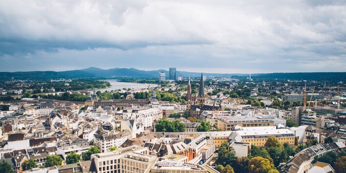 Blick vom Stadthaus Bonn. Der Blick geht hoch hinweg über Hochhäuser in die Ferne zu Wasser und einer leicht bergigen Landschaft. Das Wasser und die Landschaft liegt im blauen Dunst.