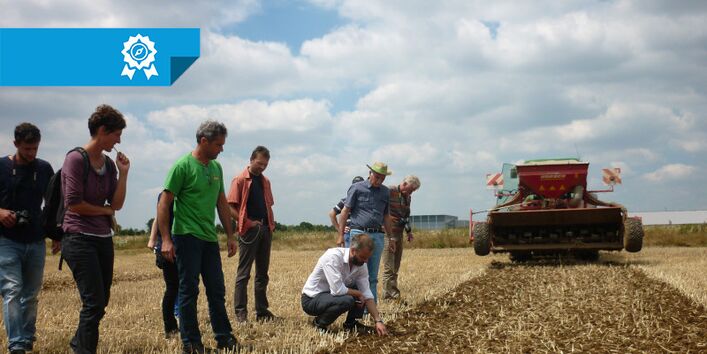 Mehrere Personen stehen auf einem Feld. Im Hintergrund fährt ein Mähdrescher. Einige der Personen begutschten den Boden.