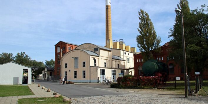Bildungs- und Transformationszentrum Brikettfabrik LOUISE. Das historische Gebäude hat eine helle Fassade und grau abgesetzte Fenster und runde Dächer. Das Gebäude steht an der geteeerten Fahrbahn an der äußeren Kurve. Es stehen mehrere Gebäude darum herum. Im Hintergrund ist der blaue Himmel sowie ein Schornstein zu sehen.