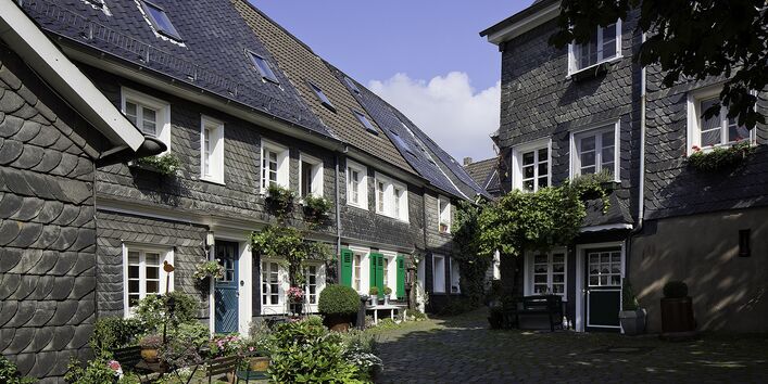 Zu sehen ist ein Ausschnit des Ortes Solingen. Zwei grauweiße Gebäude stehen schräg zu einander. Der blaue Himmel ist im Hintergrund oben zu sehen.