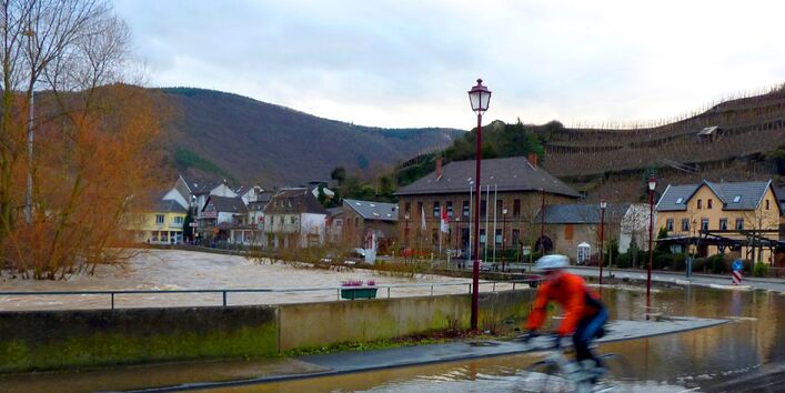 Person fährt mit dem Fahrrad durch Hochwasser in einer Ortschaft. Im Hintergrund sind Häuser und Berge zu sehen. Himmel ist bewölkt.