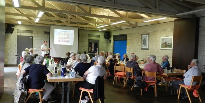 Bürger*innen sitzen im Vortragsraum an zwei langen Tischen parallel zu einander und schauen nach vorne auf die Präsentationsleinwand.