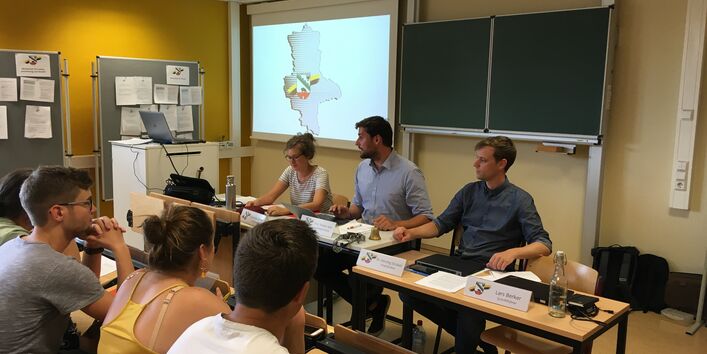 In einem kleinen Hörsaal sitzen drei Personen mit Blickrichtung in den Raum hinein. Ihnen gegenüber sind mehrere andere Personen zu sehen. Vor den Personen am Kopfende stehen Namensschilder.